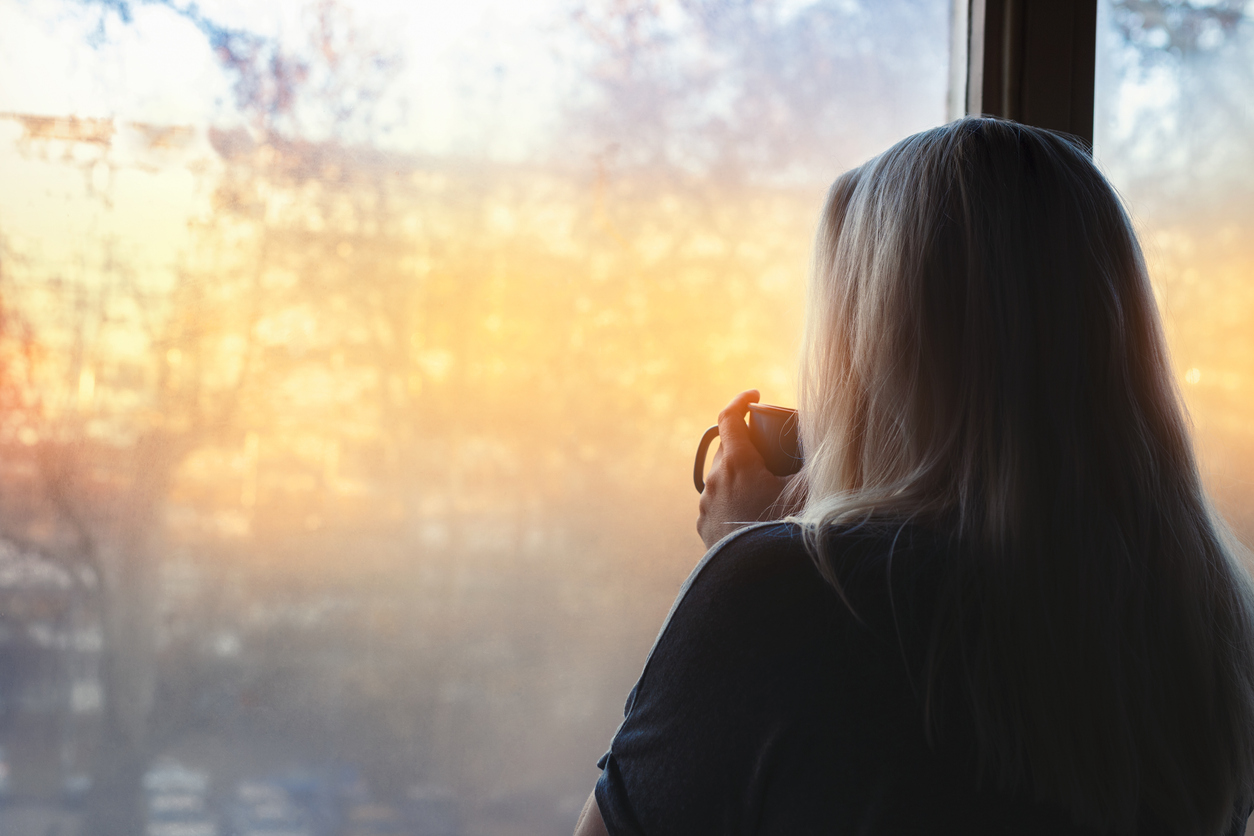Woman thoughtfully out a window, backlit by the sun