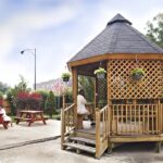 Wooden gazebo in a community yard space surrounded by picnic tables, brush, and a fence.