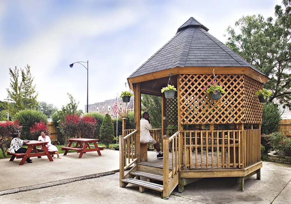 Wooden gazebo in a community yard space surrounded by picnic tables, brush, and a fence.